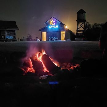 Lark Ranch - A nighttime outdoor scene shows several individuals sitting around a campfire, which is burning with visible flames and smoke. In the background, there is a rustic ranch-style building with a water tower nearby, illuminated by exterior lighting. The setting appears rural, with open space and minimal ambient light, emphasizing the glow of the fire and the illuminated building. - Ranches Near Me - Greenfield, Indiana