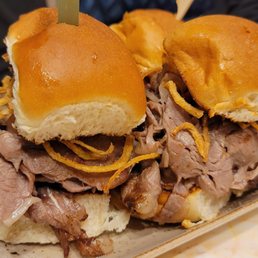 Prime Rib - Three prime rib sliders featuring shredded beef, crispy fried onions, and soft sandwich buns are arranged on a rectangular ceramic plate. The sliders are garnished with small skewers and are served in a casual dining setting, likely at Biscotti's Café in Reno, Nevada, which offers a variety of sandwiches and dining options.
