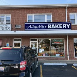 Exterior of Bakeshop - The image shows the storefront of Allegretti's Bakery in a brick building, with signage and display windows, and parked cars in the parking lot.
