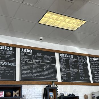 The Native Blends - Menu - A blackboard menu mounted on a white wall in a coffee shop features sections for coffee, teas, shakes, and waffles. The menu lists various drink options, flavors, and toppings, with detailed descriptions. The ceiling has a grid pattern with fluorescent lighting, and a security camera is visible in the corner. - Juice Bars & Smoothies Near Me - San Marcos, Texas