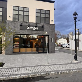 Wonder Cresskill - The image shows the exterior of Wonder, a business with a modern facade that includes large glass windows and a black sign with white lettering. The building has a two-story structure with a combination of light-colored upper walls and dark brick lower walls. The sidewalk in front is paved with gray bricks, and there is a streetlamp and a street sign nearby. The scene appears to be in an urban or suburban area with other buildings and parked cars visible in the background. - Food Delivery Services Near Me - Cresskill, New Jersey