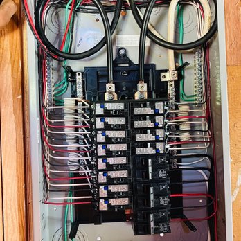 LRB Electric - Sub Panel 100 amp - The image shows an open metal electrical panel with numerous circuit breakers arranged in two columns. Inside, there are various wires, including black, red, green, and white, connected to the breakers and other terminals. The wiring is organized and secured, with some wires looping and others running along the sides. The panel is mounted on a wooden surface, and the interior components are visible, including bus bars and connection points. - Electricians Near Me