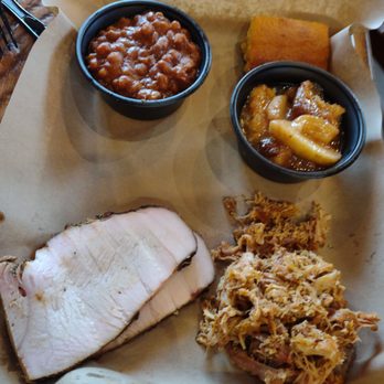 Mission BBQ - Turkey and Pulled Chicken with Baked Beans - A tray contains a meal with sliced pork, baked beans in a small black cup, macaroni and cheese in another black cup, and pulled pork. The food is served on a paper-lined tray on a wooden table, typical of a barbecue restaurant setting. - Barbeque Near Me - Springfield, Illinois