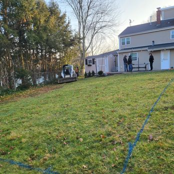 The Pool Boss - Pool Markings - A residential backyard features a multi-story house with beige siding and a deck area. Several people are gathered near the back door, engaged in conversation. The yard has a grassy lawn with blue markings and a small excavator on the left side. Tall trees and a chain-link fence border the yard, with a shed visible in the background. The scene is illuminated by natural daylight. - Pool & Hot Tub Service Near Me - Wayne, New Jersey