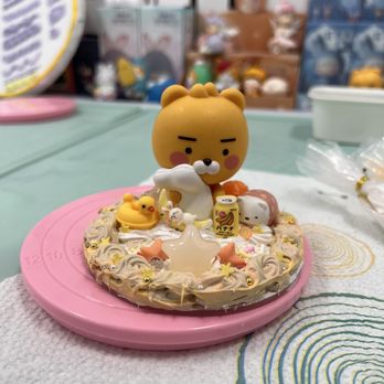 Crafts Zone Studio - Decoden Cream Phone Stand - A cake decorated with a bear figurine, small toy animals, and various decorative elements, including a small can and star-shaped decorations. The cake is placed on a pink tray on a table with a textured cloth, with a blurred background showing other toys and items. The decorations include pastel colors and small edible or plastic figures, creating a playful presentation. - Paint & Sip Near Me - Brookline, Massachusetts