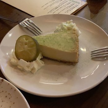 Bascule Bridge Grille - Key lime pie - A slice of key lime pie with a green topping and a graham cracker crust is served on a white plate. The plate also contains whipped cream with a pickle slice on top. The setting includes forks on either side of the plate, and the background shows a wooden table with glasses and a menu. - New American Near Me - Ashtabula, Ohio