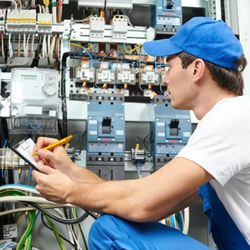 FS Electric - A technician in a blue cap and overalls kneels before a complex electrical panel, meticulously recording data on a clipboard....