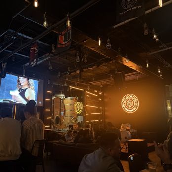 Magic 13 Brewing - An indoor brewery or bar with a stage, live music equipment, and patrons seated at tables, illuminated by warm lighting and hanging bulbs. - Breweries Near Me - Miami, Florida