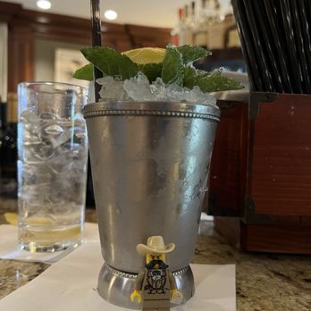 Round Robin Bar - The best mint julep we've ever had - A mint julep cocktail is presented in a metallic cup filled with crushed ice, garnished with fresh mint leaves and a lime wedge. The drink is on a bar counter, with a LEGO figure dressed as a cowboy positioned in front of the cup. In the background, there is a glass of water and bar supplies, indicating a bar setting. - Bars Near Me - Washington, DC, District of Columbia