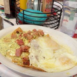 Chilaquiles - A plate of chilaquiles featuring fried eggs, diced ham, and sauce, served at Tune-Up Cafe in Santa Fe, New Mexico. The background includes condiments such as mustard and hot sauce, a glass of water, and a basket with utensils and napkins. The setting appears to be a casual dining environment with a table covered in a decorative tablecloth.