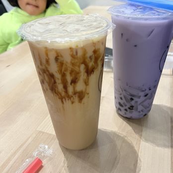 Kaito Bubble Tea - Taro Milk Tea - Two large bubble tea drinks are placed on a light wooden table inside a bubble tea shop. The drink on the left has a caramel syrup swirl, while the drink on the right is purple taro flavor with tapioca pearls at the bottom. Both drinks are in clear plastic cups with sealed lids. A pink straw is on the table. In the background, a child with curly hair wearing a neon green hoodie is slightly out of focus, raising a hand near their head. - Bubble Tea Near Me - Edison, New Jersey