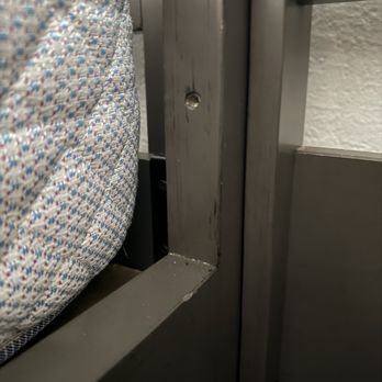 Apache Moving & Storage - A metal bed frame with a fabric-covered mattress is shown, with visible screws and a textured wall in the background. - Movers Near Me - Leander, Texas