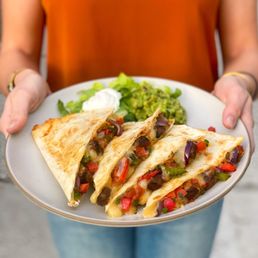 Quesadilla - A person is holding a white plate containing three grilled quesadillas filled with diced vegetables and meat, cut into triangles. The plate also includes a side of guacamole, a dollop of sour cream, and a small salad with leafy greens. The individual is wearing an orange top and blue jeans, with their arms visible in the image. The background is blurred, focusing on the food presentation.