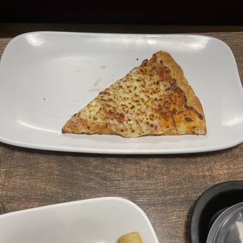 Fazoli's - Kid's cheese pizza slice. - A slice of cheese pizza with melted cheese and a slightly browned crust is positioned on a white rectangular plate. The plate is on a wooden table, with part of another plate containing fried food visible in the lower left corner. The background is dark and out of focus. - Italian Near Me - Amarillo, Texas