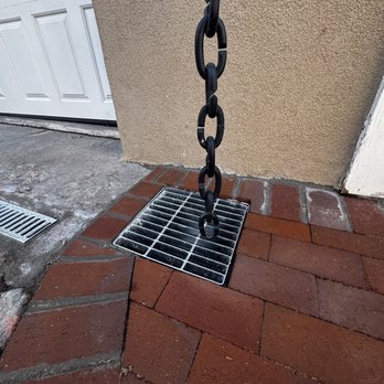 AZR Landscape & Construction - Driveway drainage and water diversion from Canales; Beautiful Brick Work - A black metal chain hangs vertically over a drainage grate on a brick patio, adjacent to a beige wall and a white garage door. - Masonry/Concrete Near Me