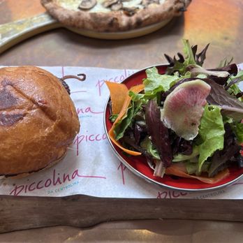 Piccolina - Polpo Panino - A sandwich with a toasted, golden-brown bun and a side salad with mixed greens and radish slices, served on a wooden board. - Italian Near Me - Washington, DC, District of Columbia