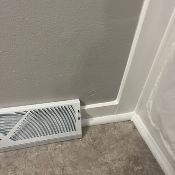 Construction Specialties - The image shows a white rectangular air vent cover with a curved grille design, positioned on the floor close to the baseboard of a gray painted wall. The wall has a textured finish, and the floor is covered with brown, stone-like tiles. A white baseboard runs along the bottom of the wall, and a plastic shower curtain is partially visible on the right side of the image. - Decks & Railing Near Me