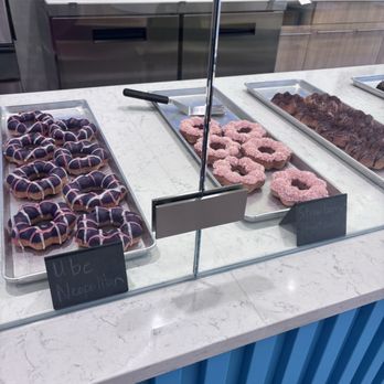 Mochinut - A bakery or cafe counter features trays of donuts and baked goods behind a glass display. The left tray contains donuts with purple icing and pink drizzle, labeled as "Ube Neopolitan." The middle tray has pink-glazed donuts with sprinkles, labeled "Strawberry Shortcake." The right tray holds chocolate or caramel glazed baked items. The counter has a white marble surface with a blue panel below, and the background shows kitchen equipment and sinks. - Hot Dogs Near Me - Columbus, Ohio