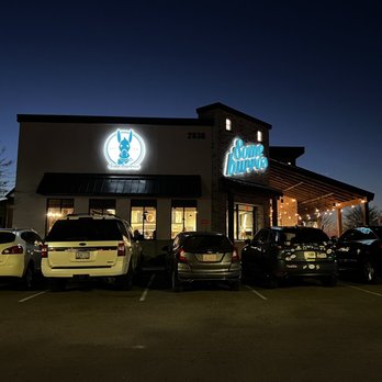 Someburros - the outside of a restaurant at night - The image depicts the exterior of Someburros restaurant located at 2836 in Mesa, AZ, taken during the evening or night. The building features illuminated signage, including a donkey logo and the Someburros name, with warm lighting under the awning. Several cars are parked in front of the restaurant, and the sky is dark, indicating nighttime. The scene is well-lit by the restaurant's exterior lights, highlighting the storefront and parking area. - Mexican Near Me - Mesa, Arizona