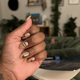 The image shows a hand with dark skin tone and decorated nails, featuring metallic gold accents and neutral beige polish, resting on a surface. The background is a blurred indoor living space with shelves, framed pictures, plants, and a person lying down, creating a cozy atmosphere.