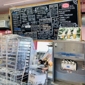 Kandy Korner - A large chalkboard menu at Kandy Korner in Hyannis, MA, features sections for homemade ice cream, soft serve, sundaes, and toppings. It lists flavors such as blueberry raspberry, cherry, kiwi strawberry, and lemon-lime, with some items marked as sold out. The menu includes details about sizes, sauces, and specialty sundaes like brownie sundae and hot fudge marshmallow. The counter below shows ice cream containers, toppings, and equipment, with a promotional photo of ice cream cones on the right. - Candy Stores Near Me - Hyannis, Massachusetts