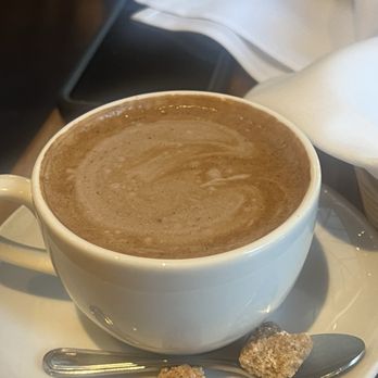 Zut On Fourth - A white ceramic cup filled with hot chocolate sits on a saucer, accompanied by two sugar cubes and a spoon. - Mediterranean Near Me - Berkeley, California