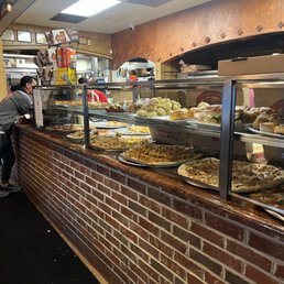 A glass display case at La Piazza restaurant contains multiple types of pizza, bread rolls, and baked dishes. The case is positioned on a brick counter, with the kitchen visible behind it. The interior features a ceiling with fluorescent lighting and a rustic decor style, including a wooden arch and wall accents. Customers are seen in the background, and the setting suggests a casual Italian dining environment.