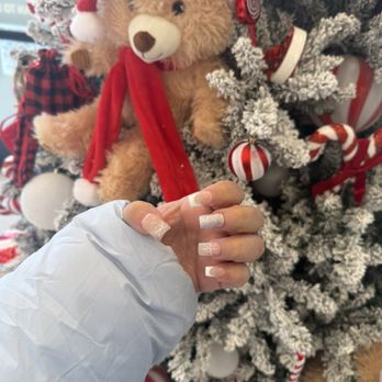 Milano Nail Spa - Done gracefully by Grace! Love my manicure! - A hand with manicured nails featuring glitter and French tips is positioned in front of a Christmas tree decorated with red and white ornaments, including a teddy bear ornament wearing a Santa hat. The person is wearing a white sleeve, and the background includes festive holiday decorations. - Nail Salons Near Me - Broadview, Illinois