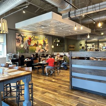 First Watch - Dining - A restaurant interior featuring wooden flooring, hanging Edison-style bulbs, and patrons seated at tables, with a mural of chickens and flowers on the wall. - Breakfast & Brunch Near Me - Newark, Delaware