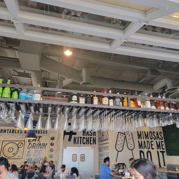 Hash Kitchen - The image shows the interior of a restaurant or bar with a ceiling-mounted shelf holding bottles of alcohol and hanging glassware. The wall has murals with text and illustrations, including phrases like 'Mimosas made me do it.' Several patrons are seated at tables, engaging in conversation and dining. The ceiling has visible ductwork and lighting fixtures, contributing to an industrial aesthetic. - Breakfast & Brunch Near Me - Chandler, Arizona