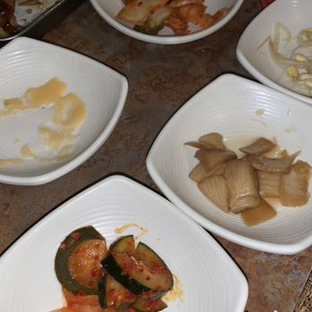 Yori - Sides - Multiple white bowls containing remnants of Korean side dishes such as cucumber kimchi, pickled vegetables, and other fermented or pickled items are arranged on a wooden table. The dishes appear partially eaten, with some sauce or liquid remaining. The table also has a metal tray with paper, a glass of water, and a red chopstick wrapper nearby, indicating a dining setting at a Korean or Asian fusion restaurant. - Korean Near Me - Hurst, Texas
