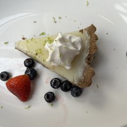 Yuzu Pie - A slice of yuzu pie with a graham cracker crust, topped with whipped cream and lime zest, served with strawberries and blueberries on a white plate.