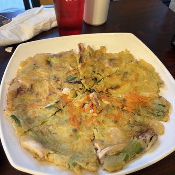 K-BAP - A Korean pancake (pajeon) with vegetables, including carrots and green onions, is placed on a white square plate on a dark wooden table. The pancake appears crispy and golden-brown, with visible vegetable pieces. In the background, there is a red Coca-Cola glass, a white condiment bottle, and a white napkin. The setting suggests a casual dining environment, likely at a Korean restaurant. - Korean Near Me - Bloomington, Indiana