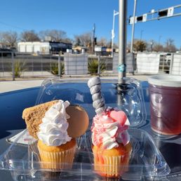 I always have to stop at A Heavenly Taste Cakery when I'm at El Vado. COFFEE CAKE & HEART SUCKER (just in time for Valentines). - Two cupcakes with decorative frosting and toppings are on a plastic container on an outdoor table. One cupcake has a cookie, whipped cream, and a swirl of frosting, while the other has pink and white frosting with a candy stick and a pink heart-shaped topping. A plastic cup with a dark beverage is next to the cupcakes. The background shows a clear blue sky, a street with some buildings, and outdoor seating areas, indicating a casual outdoor setting.