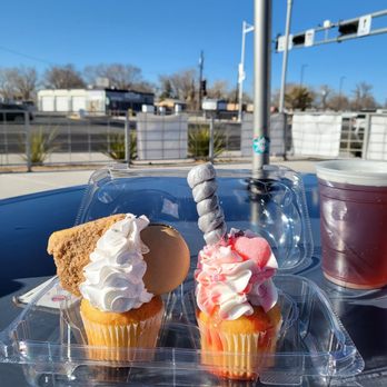 El Vado Taproom - I always have to stop at A Heavenly Taste Cakery when I'm at El Vado. COFFEE CAKE & HEART SUCKER (just in time for Valentines). - Two cupcakes with decorative frosting and toppings are on a plastic container on an outdoor table. One cupcake has a cookie, whipped cream, and a swirl of frosting, while the other has pink and white frosting with a candy stick and a pink heart-shaped topping. A plastic cup with a dark beverage is next to the cupcakes. The background shows a clear blue sky, a street with some buildings, and outdoor seating areas, indicating a casual outdoor setting. - Beer Bar Near Me - Albuquerque, New Mexico