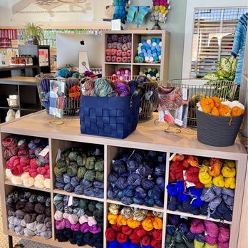 Stitch Yarn Studio - Store interior - The image shows a display of yarn skeins in multiple colors organized in cubbies and baskets at Stitch Yarn Studio. The shop features a wooden table with baskets of yarn, a shelving unit with yarn balls, and a background with knitting supplies and a computer. The shop's interior includes a sign with the business name and a window with blinds, indicating a retail environment for fiber arts enthusiasts. - Knitting Supplies Near Me - Covington, Kentucky