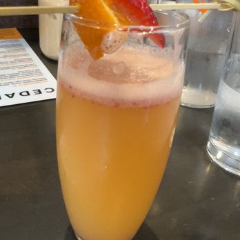 Cedar - Cedar Mosa - A tall, clear glass contains an orange-colored cocktail with a foamy pinkish top. The drink is garnished with a skewer holding a strawberry and an orange slice. The glass is placed on a dark table, with a menu, water glasses, and a jar of whipped cream visible in the background, indicating a restaurant setting. - Breakfast & Brunch Near Me - Covington, Kentucky
