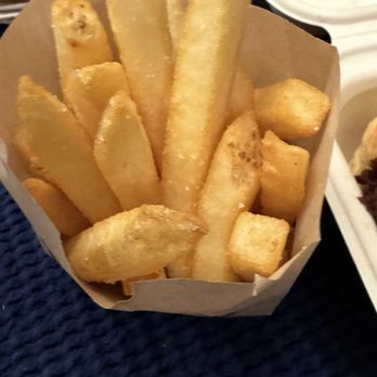 Twans Burger - A paper bag holds thick-cut French fries with a golden-brown exterior and crispy edges. The fries are arranged upright and are placed on a dark textured surface, possibly a table or tray. The background includes a white plate and part of a person's arm, indicating a dining setting, likely at a restaurant or food truck. - Food Trucks Near Me - Denver, Colorado