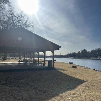 Lions Park - A lakeside park with a wooden pavilion containing picnic tables, situated on a grassy area next to a waterway. The sun is high in the sky, casting shadows from the pavilion onto the ground. Bare trees are visible in the background, along with a partly cloudy sky and distant shoreline with trees and small structures. - Parks Near Me - Fox River Grove, Illinois