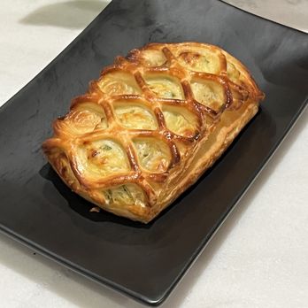 Bilqis Coffee - spinach & feta bistro - A rectangular pastry with a golden-brown, flaky crust featuring a lattice pattern on top, likely filled with cheese or vegetables. It is served on a black rectangular plate, with a white surface underneath and some green objects partially visible in the background. - Coffee & Tea Near Me - Brooklyn, New York