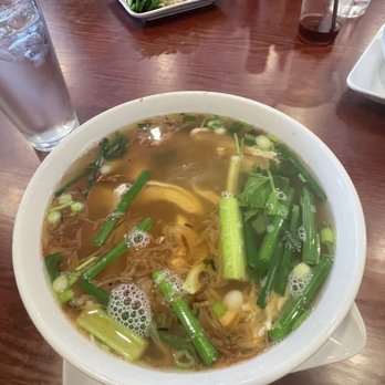 Pho One - H4. Hu Tieu Ga - A large white bowl contains Vietnamese Hu Tieu Ga soup with clear broth, green onions, leafy greens, and shredded meat. The bowl is placed on a wooden table with a glass of water nearby. In the background, there is a plate of fresh herbs and lime wedges, along with condiment bottles and a plastic cup with a straw. - Vietnamese Near Me - Metuchen, New Jersey