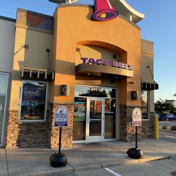 Taco Bell - The image shows the front entrance of a Taco Bell restaurant with a large, colorful bell logo mounted on the building's facade. The building has a modern architectural style with a combination of stone and stucco exterior, large windows, and a curved roof element. Two signs in the parking lot indicate designated mobile pickup parking spots, with instructions to grab inside and go. The scene is captured during daylight with a clear blue sky in the background. - Fast Food Near Me - Middleton, Wisconsin