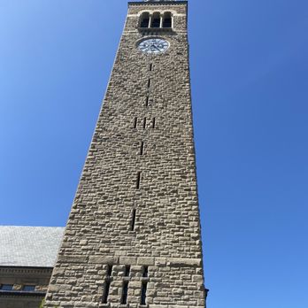 Cornell University - Clock Tower.. - A stone clock tower with a clock face near the top, set against a bright blue sky, with some greenery at the base. - Colleges & Universities Near Me - Ithaca, New York