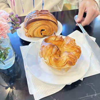 Verzenay - Delicious French food, the croissants were some of the best I've ever had! - A croissant and a pain au chocolat are placed on white plates on a black table, with a person holding a fork nearby. A small vase with flowers is also visible. - Desserts Near Me - Chicago, Illinois