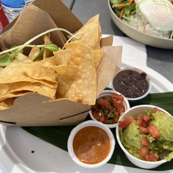 Buen Provecho - Chips - A basket of tortilla chips with side salsas and guacamole is placed on a white tray with a green leaf. In the background, a salad with a poached egg is visible. - Latin American Near Me - Albuquerque, New Mexico
