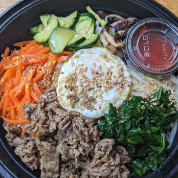 Sue's Korean Kitchen - Bibimbap - A Korean bibimbap dish presented in a black bowl includes sliced cucumbers, shredded carrots, cooked spinach, seasoned beef, and a fried egg topped with sesame seeds. A small container of sauce is also included. The ingredients are arranged separately around the bowl, showcasing traditional Korean flavors and presentation. - Korean Near Me - Stratham, New Hampshire