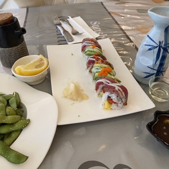 Sakura 15 - Angel Roll with Edamame & sake - A dining table features a rectangular white plate with assorted sushi rolls topped with fish, avocado, and orange fish roe. Nearby, there is a bowl of green edamame, lemon wedges, and small black dishes with dipping sauces. The table also has a ceramic sake bottle, a small glass of sake, and a black container with a cork lid. Silverware and a napkin are placed at the top of the table, and the tablecloth is covered with a plastic cover. - Japanese Near Me - Bloomington, Indiana
