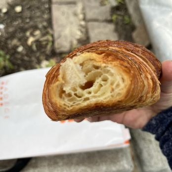 Nadege - Croissant - A hand holds a croissant showing its flaky, golden-brown crust and airy, layered interior, with an outdoor background. - Desserts Near Me - Toronto, Ontario