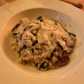 Daedalus Restaurant - Mushroom Risotto - A bowl of mushroom risotto featuring cooked rice, sliced mushrooms, and herbs, topped with shaved cheese. The dish is presented in a white, wide-rimmed bowl on a wooden table, with a glass of water and a fork nearby, indicating a dining setting. - Pubs Near Me - Cambridge, Massachusetts