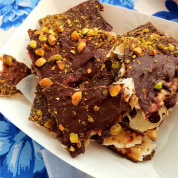 Ruthie's Bagels - Matza Crack - A serving of dessert pieces coated in chocolate with chopped pistachios sprinkled on top, placed in a white paper tray. The background features a blue and white patterned surface, suggesting a casual dining or snack setting. - Bagels Near Me - Albuquerque, New Mexico